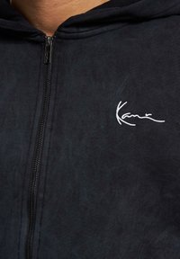 Sort zip-up hoodie lavet af blødt stof, med en tekstureret overflade og et hvidt broderet logo på venstre bryst.