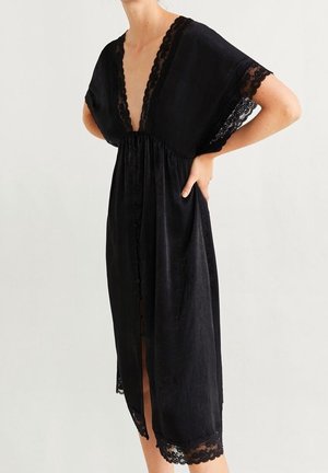 Frau trägt ein schwarzes, knöpfbares Midikleid mit Spitzenbesatz an tiefem V-Ausschnitt, Ärmeln und Saum, steht mit einer Hand in der Hüfte.