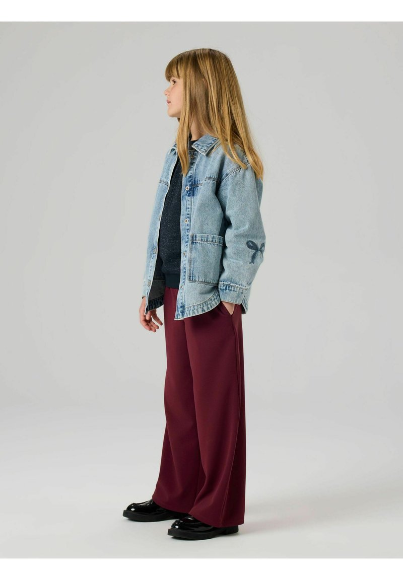Vieglā zilā krāsā džinsa jaka ar divām priekšējām kabatām, kombinēta ar platām bordo biksēm un melnām spīdīgām kurpēm. Minimālistisks dizains, stāvoša poza.