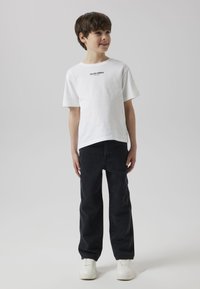 Bela bombažna majica s potiskom "Studio edition" v črni barvi, kombinirana s temnimi kavbojkami in belimi supergami, z rahlo sproščenim krojem in preprostim dizajnom.