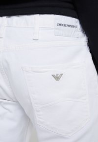 Vita denimjeans med Emporio Armani-logotyp på midjan och metallogotyp på bakfickan, kombinerat med en svart topp.
