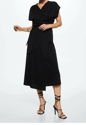 Femme portant une robe midi portefeuille noire à manches courtes et des sandales à talons hauts à lanières, exhibant son avant-bras et ses doigts tatoués.