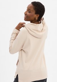 Sudadera con capucha beige, de manga larga, con una textura suave y aberturas laterales. Presenta un corte relajado, costuras visibles y capucha ajustable.
