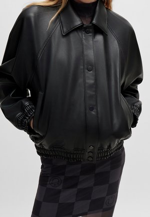 Veste en simili cuir noire avec boutons-pression, poignets et ourlet élastiques, portée sur une jupe noire à carreaux avec des visages souriants.