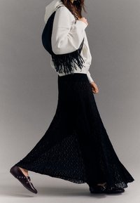 Gonna lunga nera in pizzo con texture trasparente, abbinata a un hoodie bianco. Gli accessori includono una borsa frangiata nera e scarpe flat marroni scure.