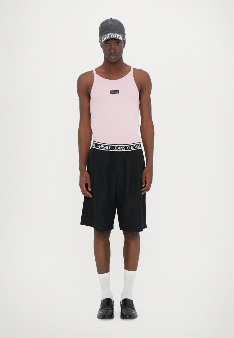 Rosa ribbet tanktopp med en logo-patch, svarte plisserte shorts med merkebånd, hvite sokker, og svarte slip-on sko.