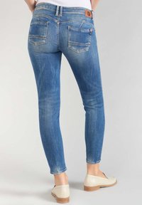 Jeans de denim azul ajustados con un acabado desgastado, presentan bolsillos traseros y un parche de cuero marrón con el logo en la cinturilla.