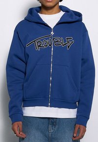 Niebieska bluza z kapturem zapinana na zamek, wykonana z mieszanki bawełny. Ma na sobie wyraziste czarne litery "TROUBLE", kieszenie kangurze oraz ściągacze na rękawach i u dołu.