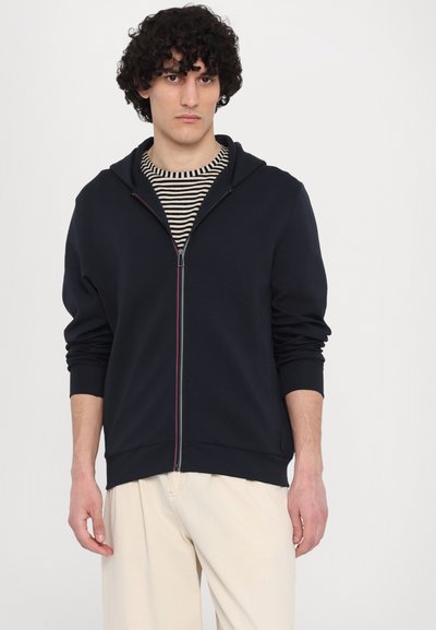 Sudadera con cremallera en color azul marino, fabricada en tela suave, con cremallera frontal y detalles en varios colores, además de una capucha. Lleva puesta sobre una camiseta a rayas.