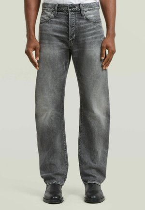 Man draagt loszittende, vervaagde zwarte jeans en zwarte leren schoenen, staand tegen een effen lichte achtergrond.