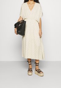 Frau in einem beige gepunkteten Midikleid, schwarzen Schnür-Plateau-Sandalen und mit einer großen schwarzen geflochtenen Handtasche.