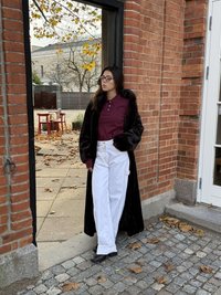 Palton lung de blană eco neagră, tricou polo burgundy, pantaloni albi cu picioare largi și pantofi negri. Fundal cu perete de cărămidă și frunze căzute.