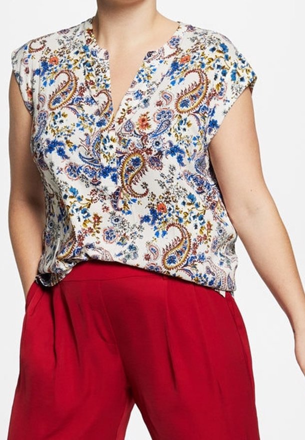 Femme portant un chemisier blanc sans manches avec des motifs floraux et cachemire bleus, rouges et bruns, associé à un pantalon rouge taille haute.
