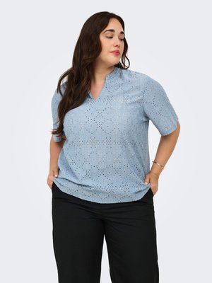 Woman with long dark hair wearing a light blue short-sleeve eyelet blouse and black pants, standing with hands in pockets, looking sideways.