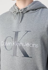 Primer plano de una persona que lleva una sudadera gris con el logo de "Calvin Klein Jeans" en blanco y las grandes letras superpuestas "CK" en el pecho.
