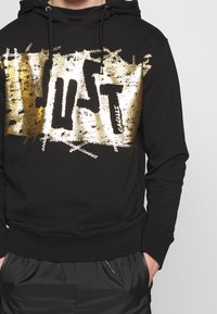 Černá mikina s kapucí s velkým zlatobílým nápisem "JUST" přes přední část, vybavená kapucí se stahovací šňůrkou a žebrovanými lemy na rukávech.