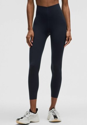 Person iført sorte leggings med høj talje og hvide atletiksko, stående mod en ensartet lys baggrund.