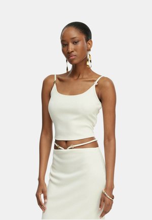 Donna che indossa un crop top bianco a coste senza maniche e una gonna abbinata con spalline sottili e accessori dorati, posando su uno sfondo semplice.