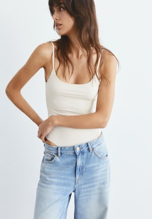 Vrouw met lang bruin haar, een wit mouwloos topje en lichtblauwe jeans met hoge taille, staand met de armen over elkaar ter hoogte van de taille.