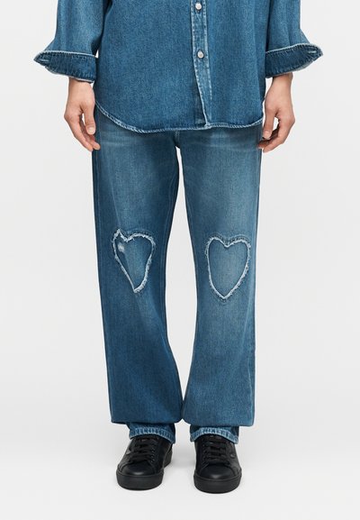 Άτομο που φορά μπλε τζιν με φθορές σε σχήμα καρδιάς στα γόνατα, συνδυασμένο με τζιν πουκάμισο στο ίδιο χρώμα και μαύρα αθλητικά παπούτσια.