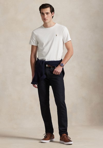 Young man wearing white short-sleeve shirt, dark jeans, brown shoes, blue watch, with a navy sweater tied around his waist, standing against beige background.