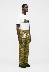 Λευκό βαμβακερό μπλουζάκι με μαύρο και καμουφλάζ σχέδιο, σε συνδυασμό με παντελόνι καμουφλάζ πράσινου χρώματος με λευκό κεντημένο μοτίβο και σκούρα πράσινα παπούτσια.