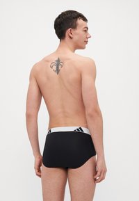 Melni peldkostīmi ar baltu elastīgu jostu, uz kuras ir trīs melnas svītras. Modeles augšējā muguras daļā ir redzams tetovējums.