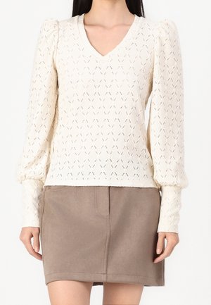 Kobieta w kremowej, teksturowanej bluzce na długi rękaw z bufkami na ramionach oraz taupe spódnicy do kolan.