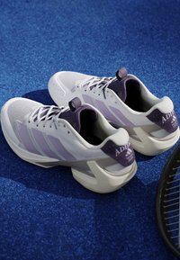 Paire de chaussures de tennis Adidas blanches et violettes placées sur une surface de court de tennis bleue à côté d'une raquette de tennis.