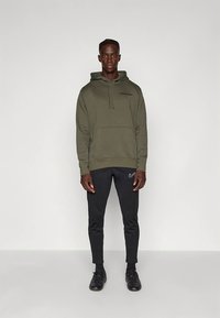 Zöld kapucnis pulóver "PARIS" felirattal és Nike logóval, fekete melegítőnadrággal és fekete sportcipővel párosítva. Simább tapintású szövet, hétköznapi kialakítás.