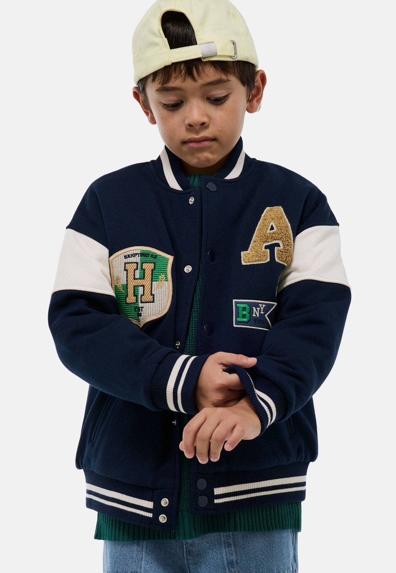 Blouson universitaire bleu marine avec accents blancs, présentant des patchs brodés, une lettre "A" texturée et des poignets côtelés. Porté par-dessus un tricot vert.