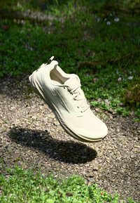 Beige Laufschuh mit strukturiertem Obermaterial, Schnürsenkeln und einem Ziehschlauf, schwebend über einem Kiesweg, umgeben von grüner Vegetation.