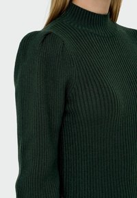 Femme portant un pull en maille côtelée vert foncé avec un col montant et des détails bouffants aux épaules, coupé au niveau du cou et de la taille.