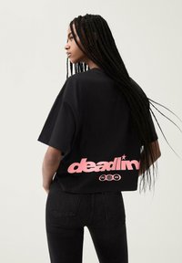 Černé bavlněné tričko s krátkým střihem, na zadní straně s růžovým logem a grafikou. Modelka má na sobě tmavé džíny. Viditelné dlouhé copy.