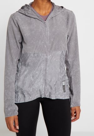 Persoon die een lichtgrijs zip-up capuchonjack met een patroonachtige textuur en een zwarte broek draagt, staand tegen een effen lichte achtergrond.