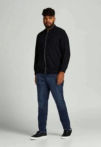 Casaco bomber preto com fecho zip e gola alta, combinado com jeans azuis e ténis pretos. Tecido suave e design ajustado.