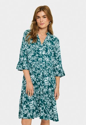 Frau trägt ein türkisfarbenes knielanges Kleid mit weißen floralen Mustern, drei Viertel langen Ärmeln und einem V-Ausschnitt, posierend vor einem schlichten Hintergrund.
