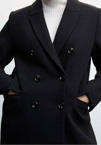 Persoon in een zwart dubbelzijdig jasje met zes knopen, handen in de zijzakken, en een witte geribbelde turtleneck trui eronder.