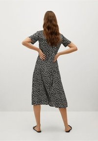 Vestido negro midi con pequeños patrones florales blancos, mangas cortas, cintura ceñida y falda fluida. Lleva sandalias planas sencillas.