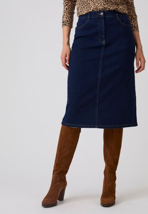 Vrouw met een donkerblauwe denim midi-rok gecombineerd met bruine suède kniehoge laarzen met hak en een top met lange mouwen en luipaardprint.