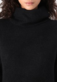 Pull à col roulé noir en tissu côtelé et texturé. Présente un col haut et une coupe décontractée. Idéal pour superposer ou porter seul.