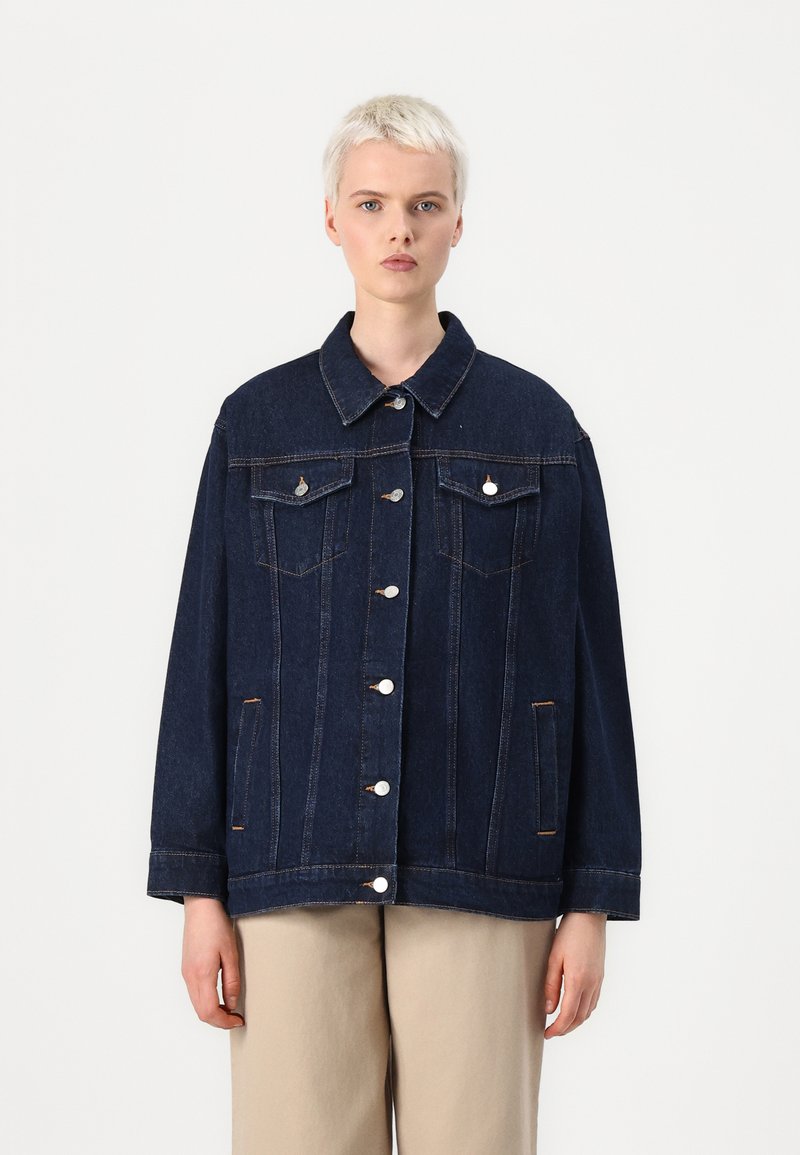 Temno modra jeans jakna z gumbastim zapiranjem spredaj, dvema prsnikoma in ovratnikom. Ima srebrno kovinsko opremo in vidne detajle šivanja.