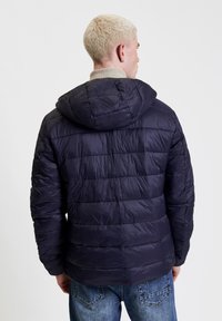 Giacca in blu navy imbottita con cappuccio, caratterizzata da un design trapuntato e un tessuto testurizzato. Il colletto è di un beige chiaro.