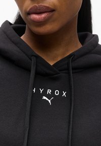 Κοντινό πλάνο ατόμου που φοράει μαύρη μπλούζα με κουκούλα, με τα λογότυπα "HYROX" και Puma κεντημένα στα λευκά στο στήθος.