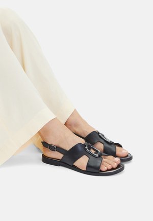 Czarne skórzane sandały o otwartej konstrukcji, z regulowanym paskiem na klamrę i ozdobnym metalowym akcentem, zestawione z jasnymi spodniami.