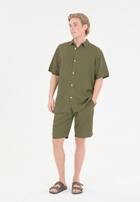 Oliivinvihreä lyhythihainen paita napilla, yhdistettynä samanvärisiin shortseihin ja ruskeisiin sandaaleihin. Silkkinen kangas, rento malli ja taskut.