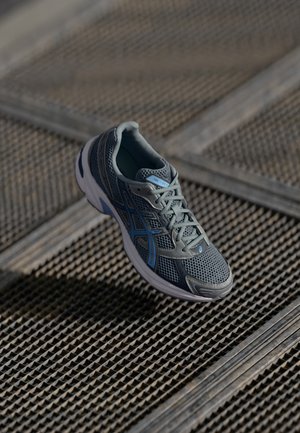 Baskets athlétiques grises et bleues en mesh et matériaux synthétiques reposant sur une surface en grille métallique texturée.