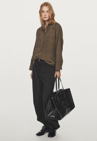 Haut en suede marron avec un col, pantalon noir ample, et un sac fourre-tout noir texturé avec deux poignées. Des chaussures noires complètent l'ensemble.