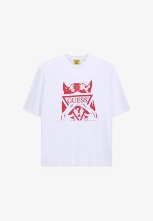 T-shirt blanc avec un graphisme rouge d'une personne avec des cornes de diable tenant un triangle avec le mot "GUESS" et un point d'interrogation à l'intérieur.