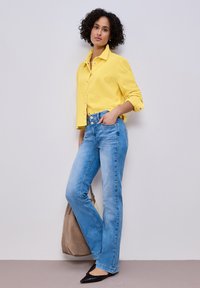 Femme aux cheveux bouclés portant une chemise jaune boutonnée, un jean taille haute bleu clair, des chaussures noires à bout pointu, et tenant un sac fourre-tout beige.